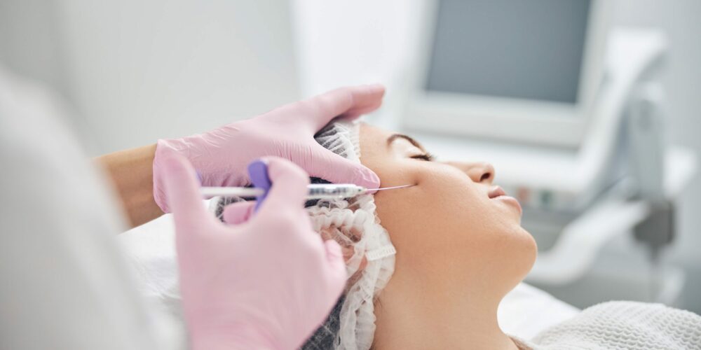 Beauty specialist with gloved hands holding a syringe and applying rejuvenation liquid in female cheek